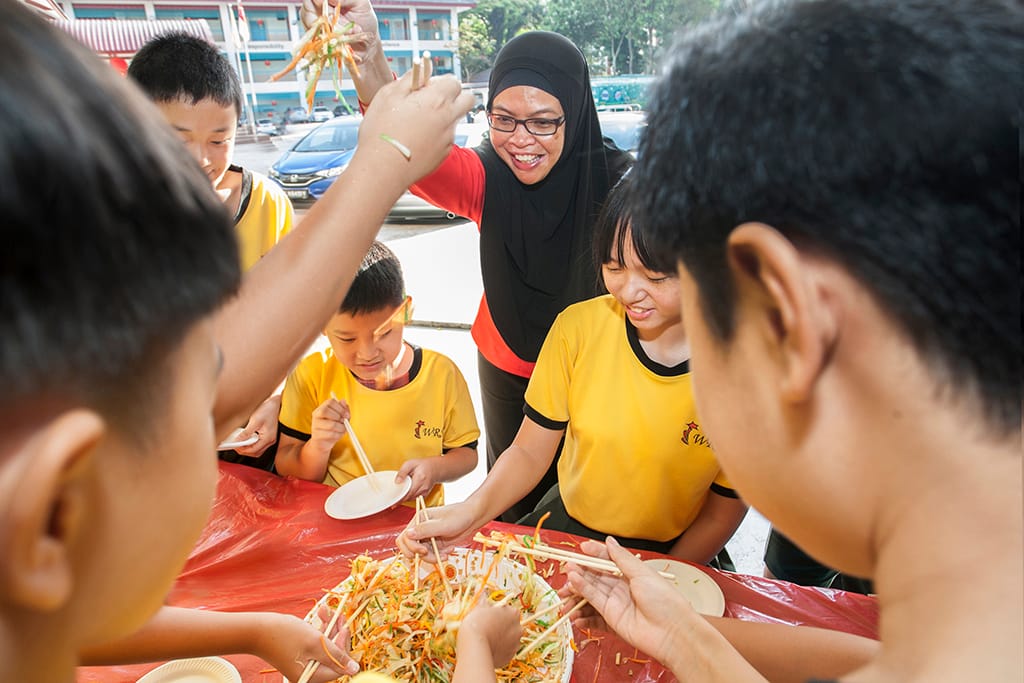 A smiling woman in a hijab and children in yellow shirts toss a colorful salad with chopsticks at a red table.